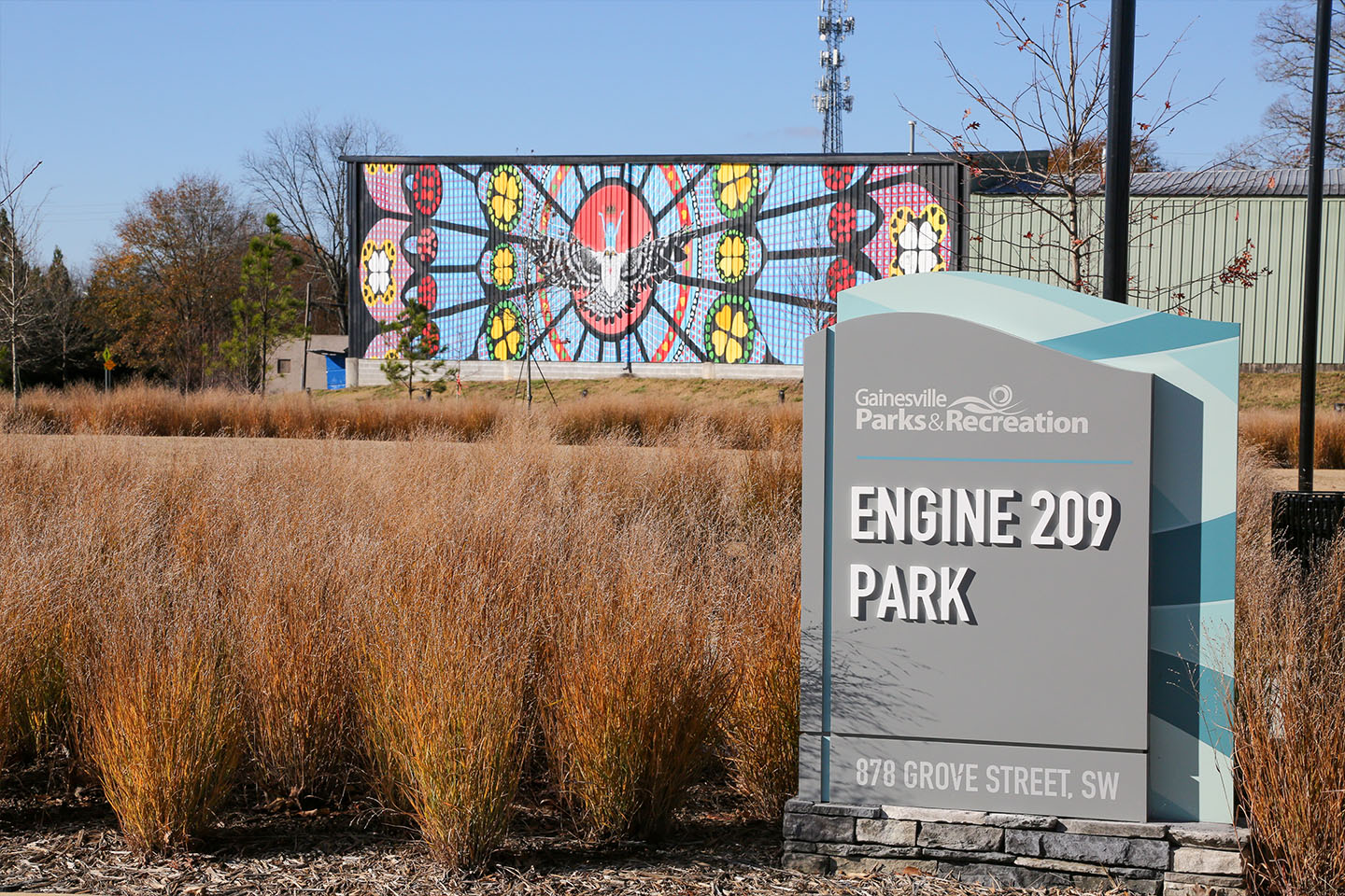a sign for Engine 209 Park, Gainesville Parks and Recreation, 878 Grove Street, SW - in the background is a mural that looks like a kaleidoscope pattern with a large bird in the center