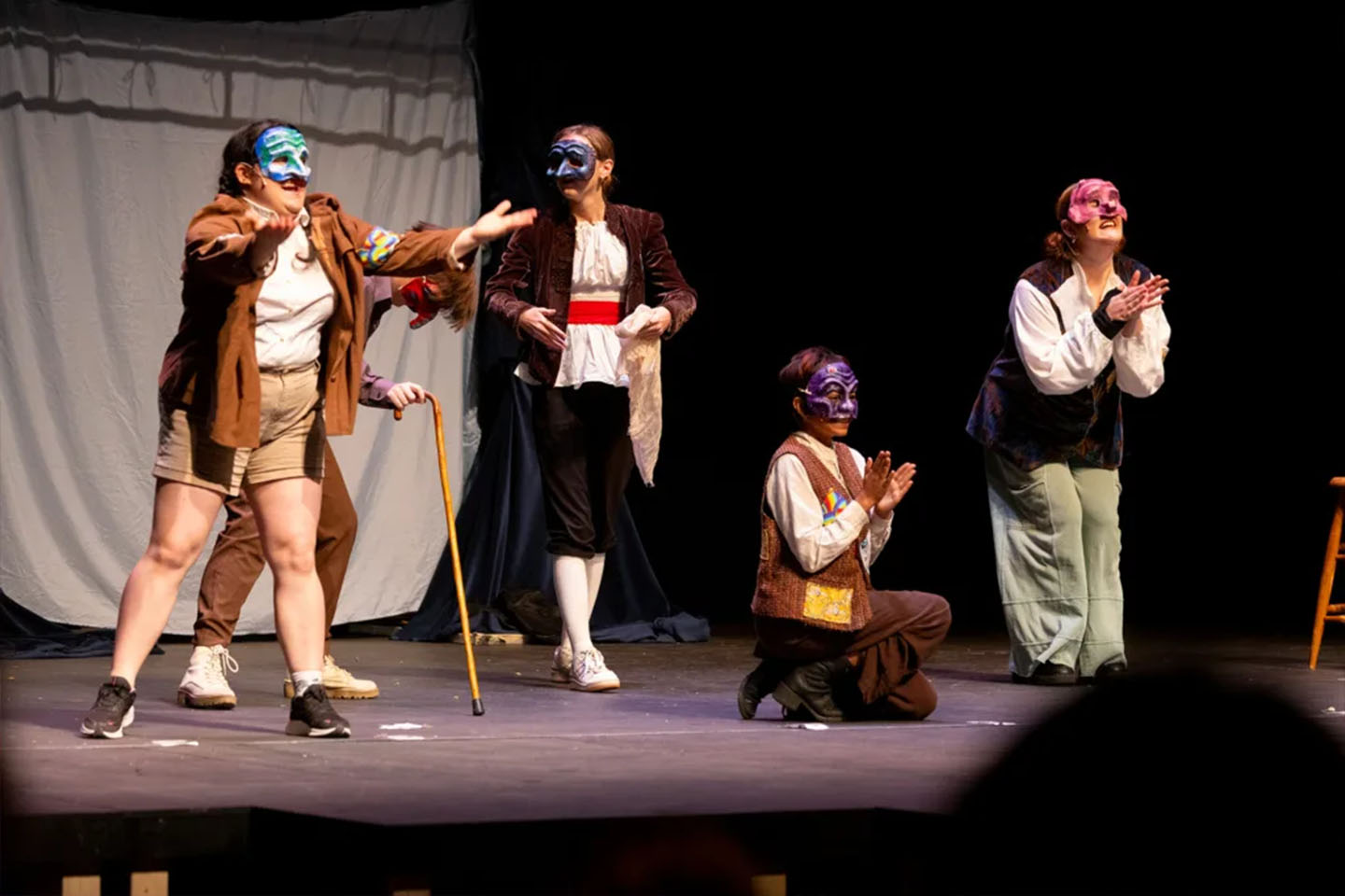 Masked student performers gesture dramatically onstage, some kneeling and clapping during lively scene.