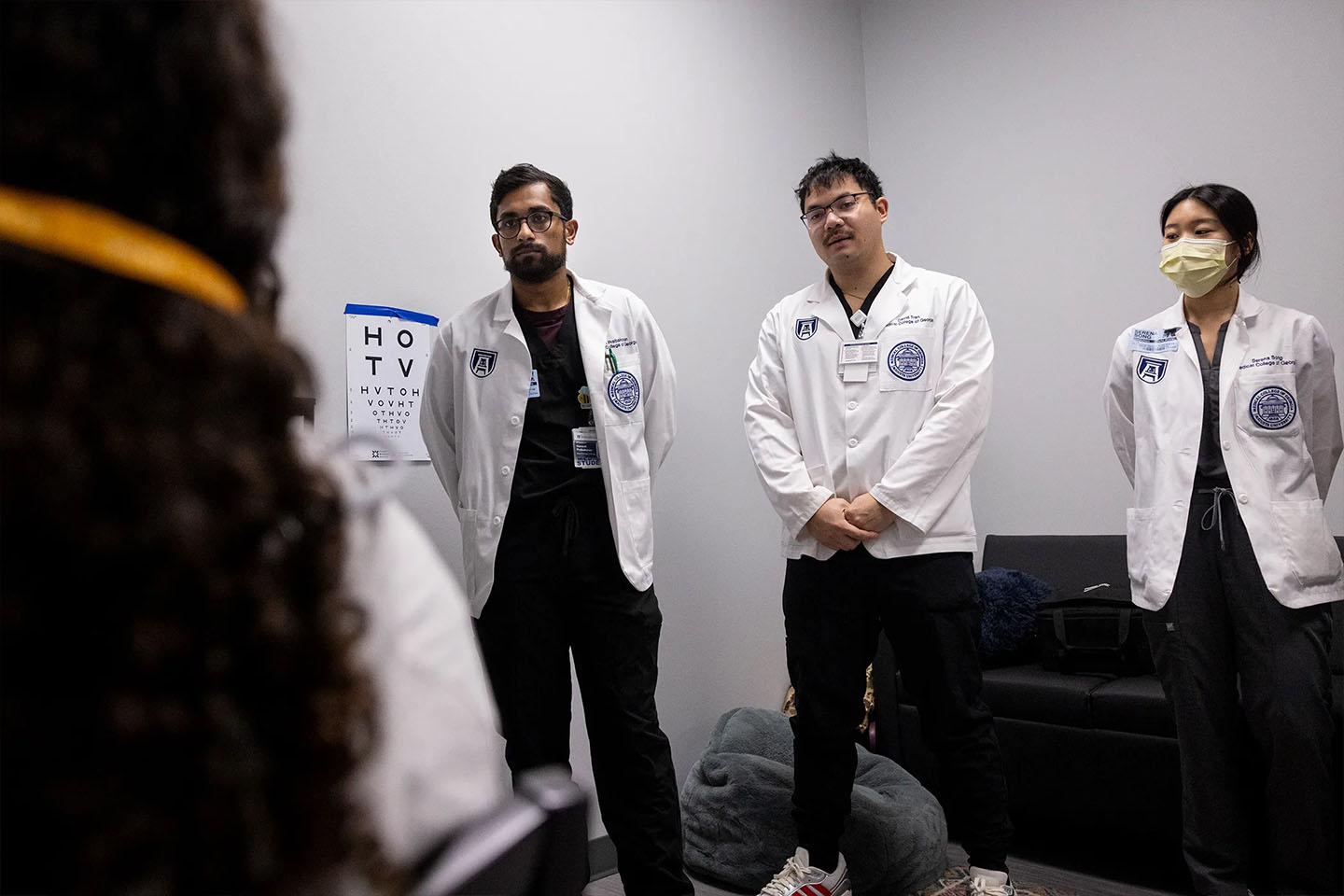 Medical students in white coats observe vision screenings inside school health center room.